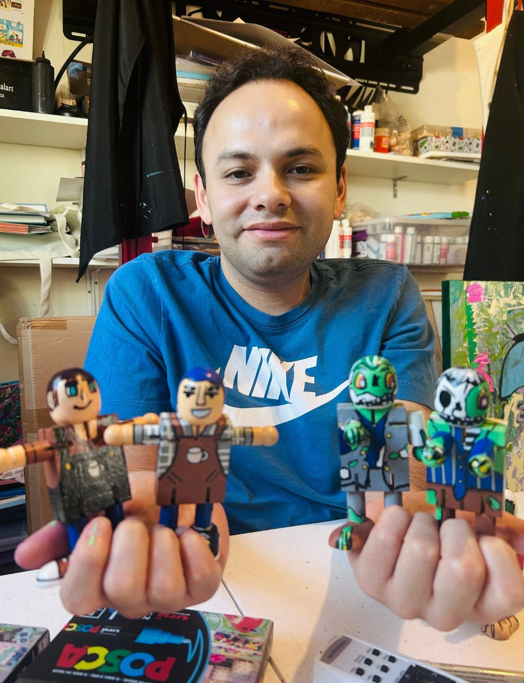 A man wearing a blue Nike t-shirt holds up four small action figures in both hands, with shelves and household items visible in the background.